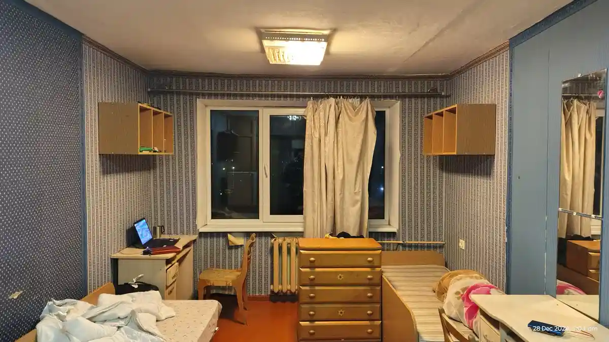 A narrow, cluttered bedroom with two beds, a wooden dresser, and a desk with a laptop, all under a single ceiling light. The…