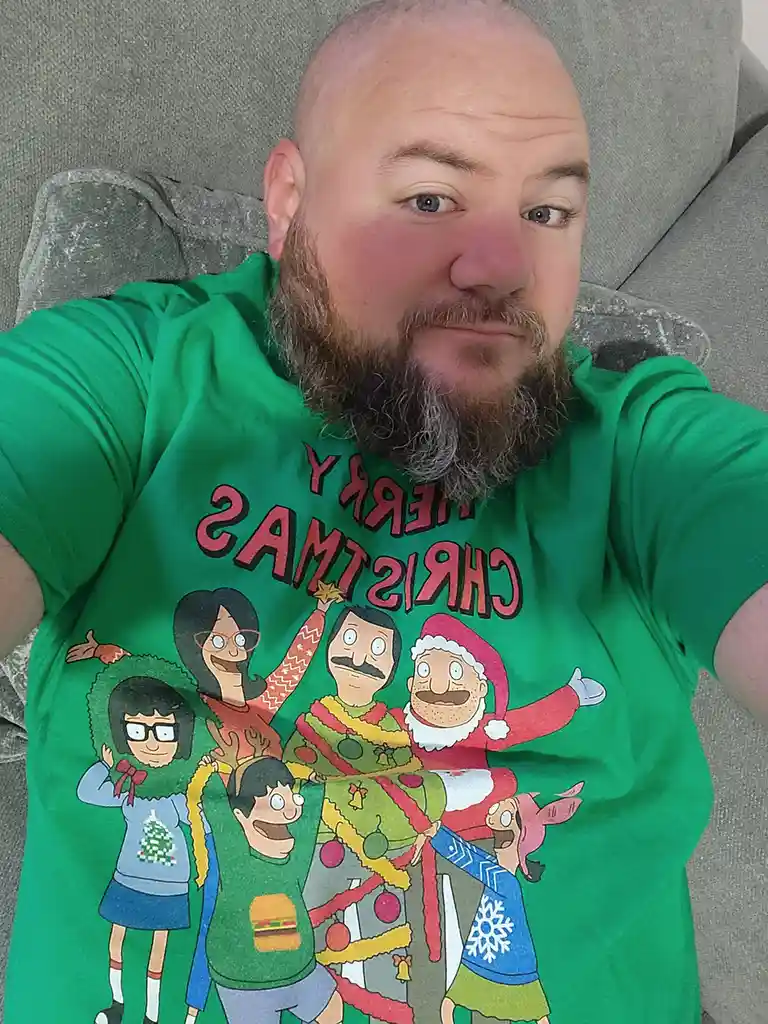 A man with a beard and shaved head lies on a grey couch wearing a green t-shirt featuring cartoon characters from 'Bob's…