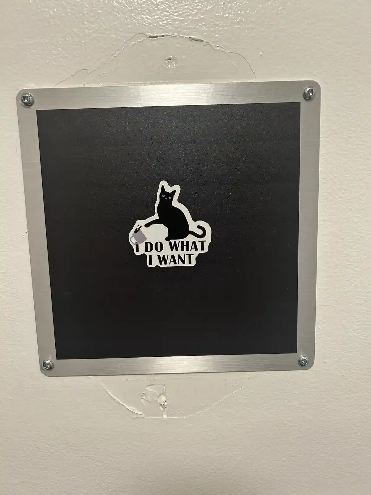 A black sticker on a metal-framed sign mounted on a wall shows a black cat holding a mug, with the text 'I DO WHAT I WANT'…