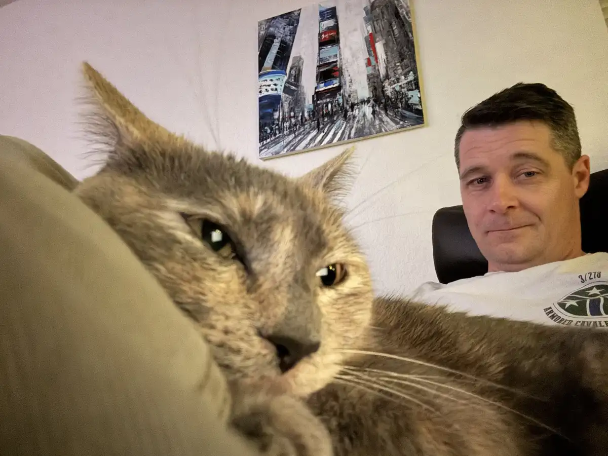 A close-up selfie shows a grey tabby cat in the foreground with one eye half-closed and a smug expression, while a man in a…