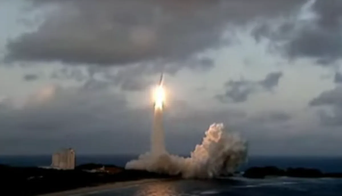 A rocket launches from an ocean-side launchpad, leaving a bright trail of fire and smoke against a cloudy sky. The overlaid…