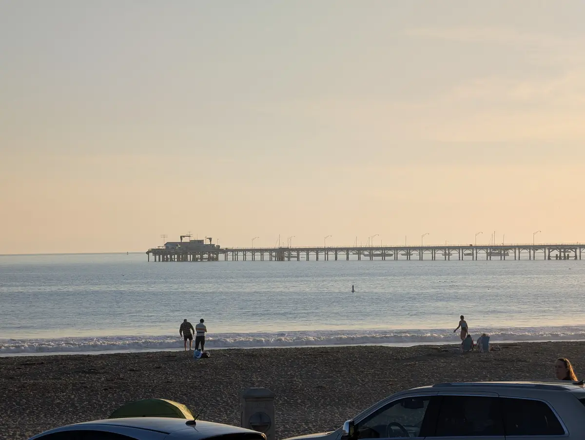 A serene beach scene at sunset with a long pier stretching into the ocean, people relaxing on the sand, and cars parked…