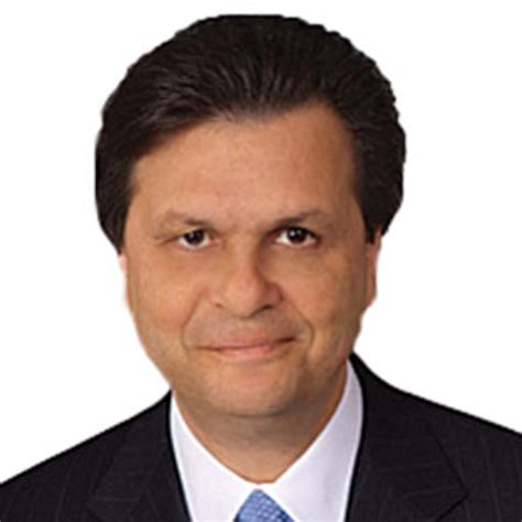 A man in a dark suit and blue tie with a slight smile looks directly at the camera against a white background. The text…