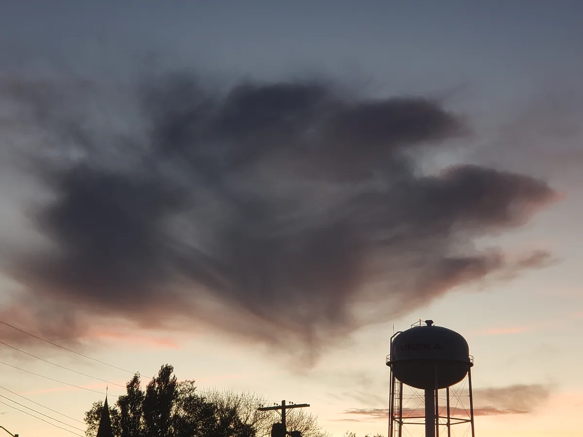 A silhouette of a water tower labeled 'EUREKA' stands against a dramatic sky at dusk, with large, dark clouds swirling above…
