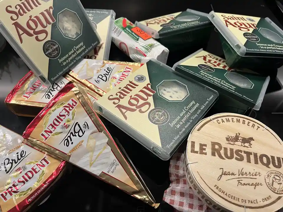 A pile of assorted French cheeses including Saint Agur blue cheese, President Brie, and Le Rustique Camembert are displayed…