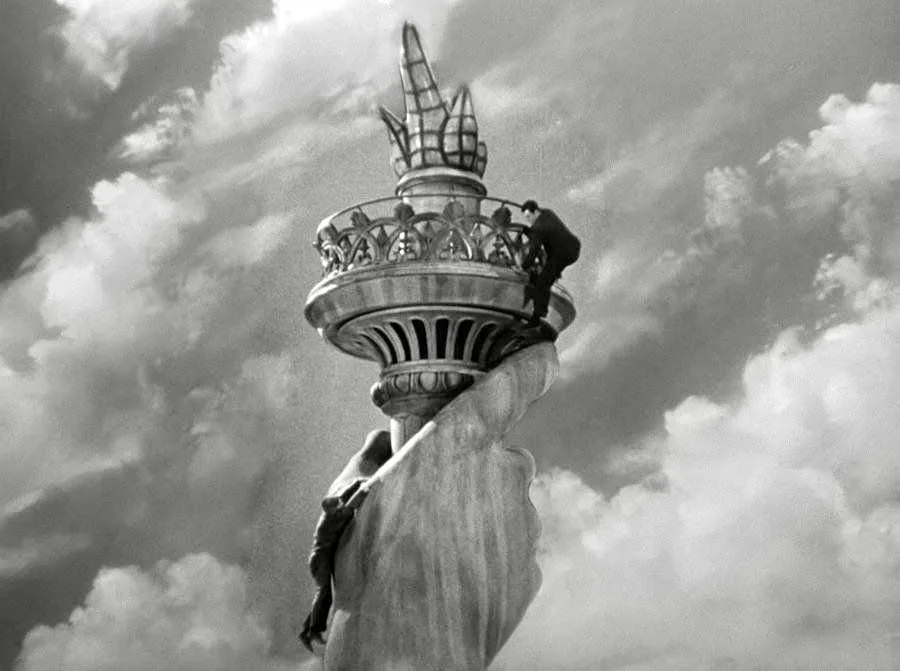 A black-and-white photo shows a person clinging to the torch of the Statue of Liberty, appearing to be stuck or stranded on…