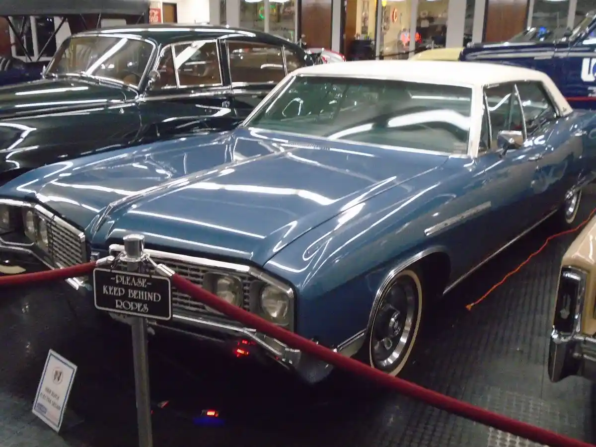 A blue and white classic car is displayed in a museum, cordoned off by red ropes and a sign that reads 'PLEASE KEEP BEHIND…