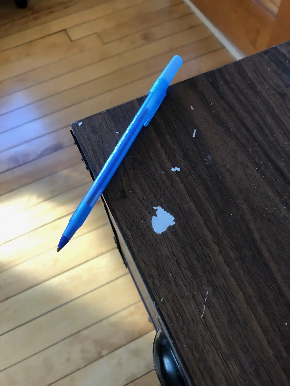 A blue ballpoint pen rests diagonally on a dark wooden table with visible scratches and a white paint chip. The pen's tip…