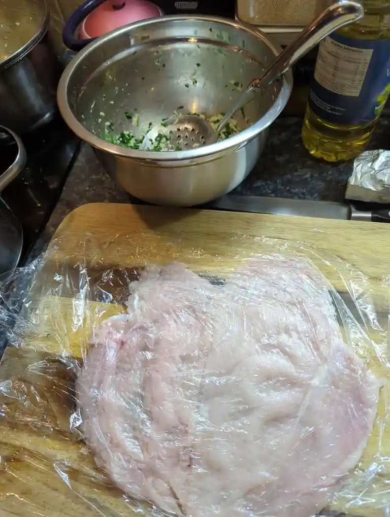 A raw, pale pink fish fillet is covered in plastic wrap on a wooden cutting board, with a metal bowl containing chopped…