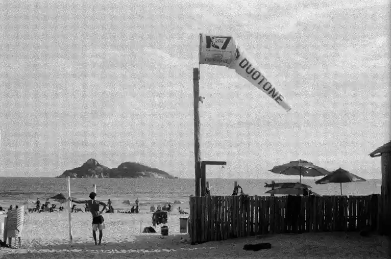 A grainy black-and-white photo of a beach scene with a man standing near a volleyball net, a wooden fence, beach umbrellas…