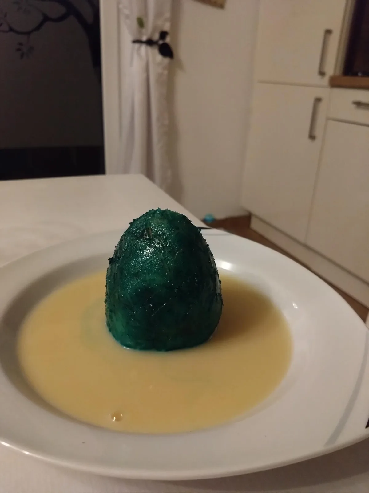 A close-up photo shows a dark green, dome-shaped food item sitting in a pool of pale yellow sauce on a white plate. The…