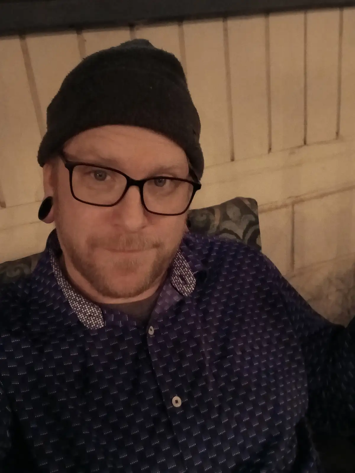 A man with glasses, a black beanie, and a large black ear gauge looks directly at the camera with a slight smirk while…