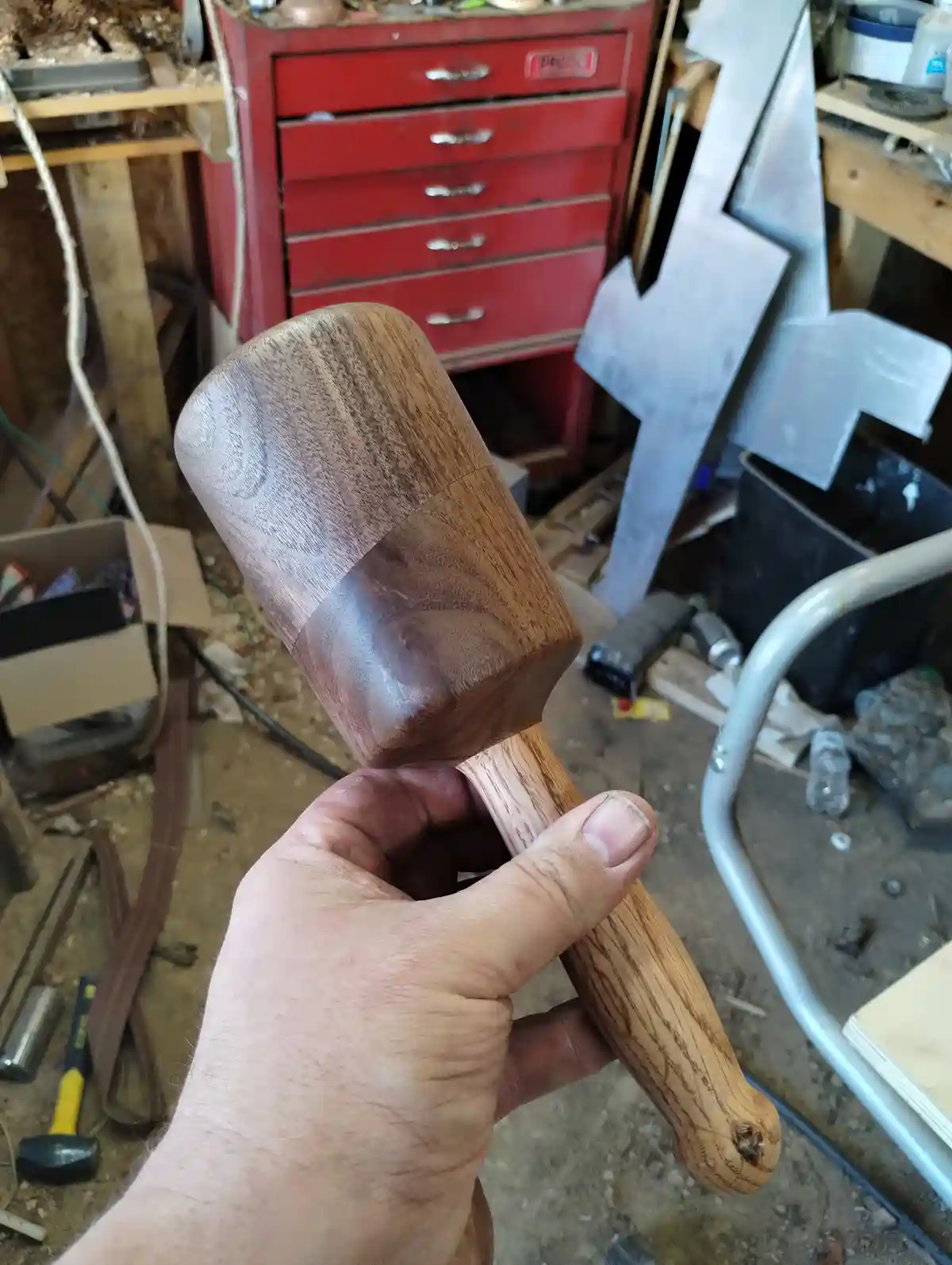 A hand holds a custom-made wooden hammer with a polished head and handle, showcasing craftsmanship in a cluttered workshop…