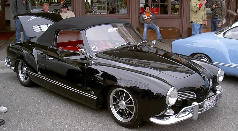 A glossy black vintage convertible with a red interior and chrome accents is parked outdoors, its hood open. Several people…