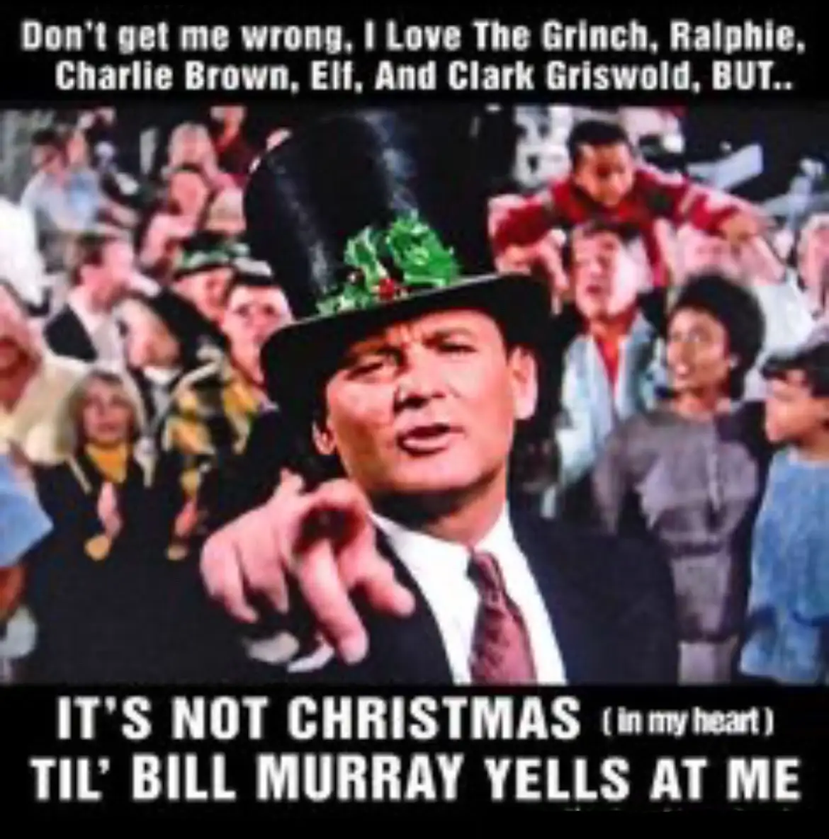 A still image of Bill Murray in a black top hat decorated with green holly, pointing forward with a stern expression. He is…