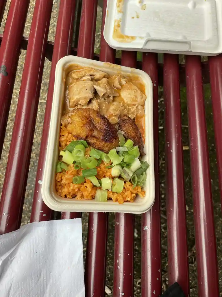 A top-down photo of a takeout container on a red metal bench, filled with orange rice, chicken in sauce, fried plantains…