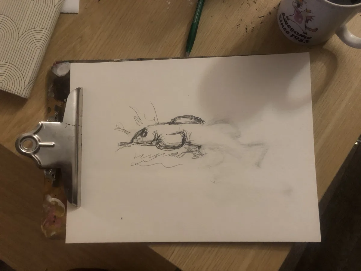 A rough pencil sketch of a fish on a white sheet of paper clipped to a metal clipboard, resting on a wooden desk. The…