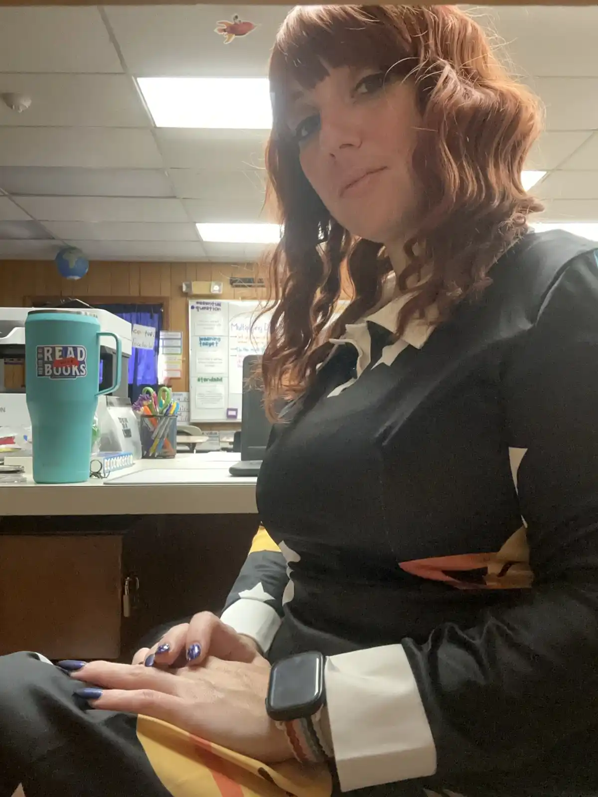 A woman with reddish-brown hair and a stern expression sits in what appears to be a classroom or office, wearing a black…