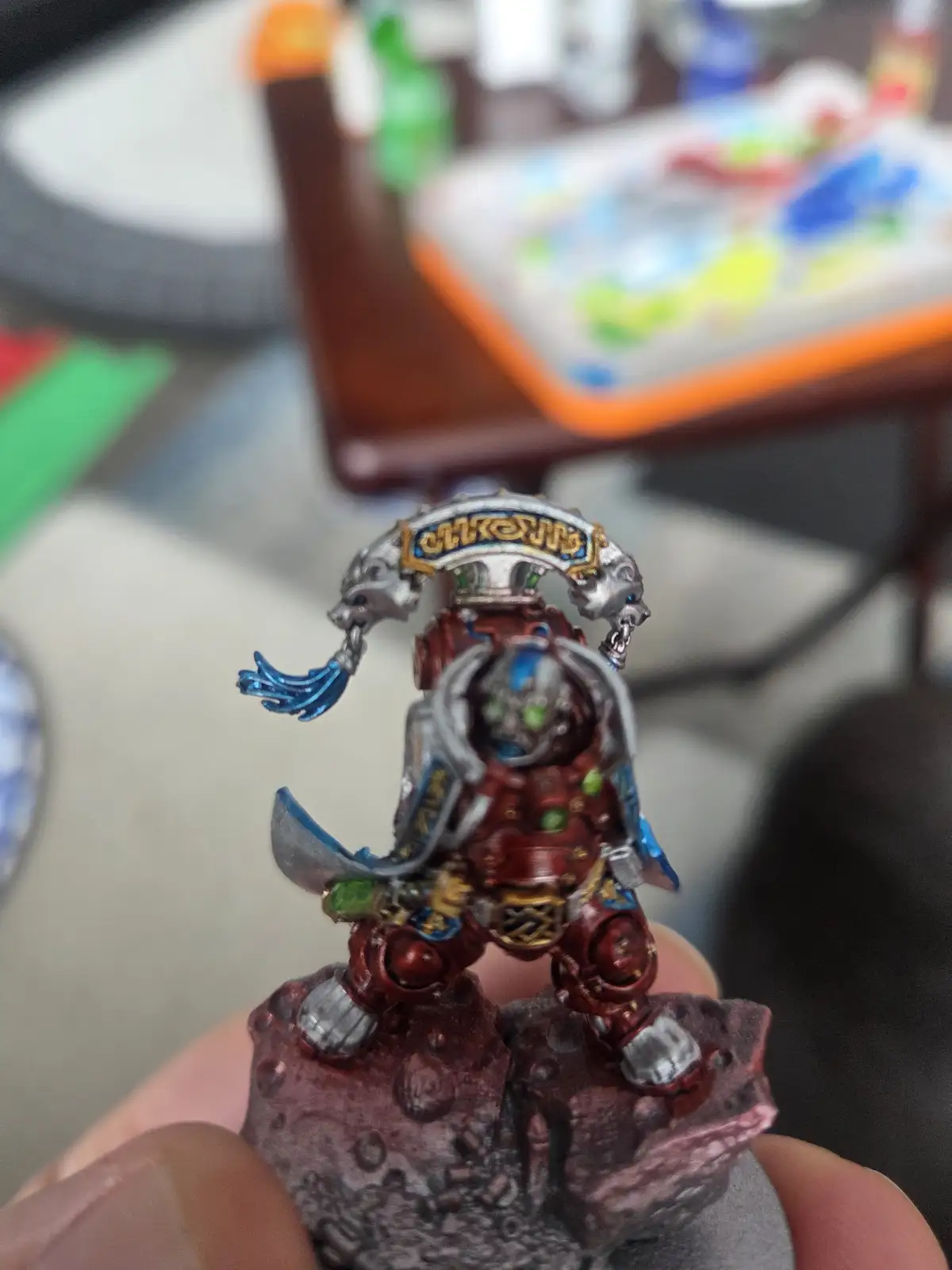 A close-up photo shows a hand holding a highly detailed, custom-painted miniature figure of a red-armored warrior with a…