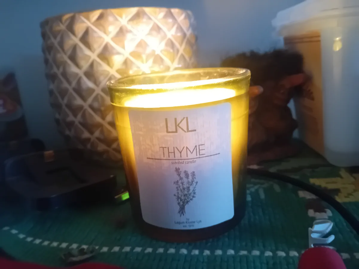 A lit, amber-colored candle in a glass jar with a white label reading 'LKL THYME scented candle' and an illustration of…
