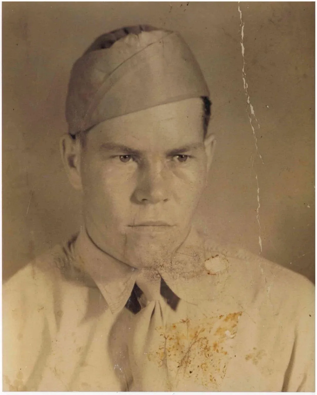 A sepia-toned, vintage, slightly damaged photograph of a man in a military-style cap and uniform shirt, looking intensely at…