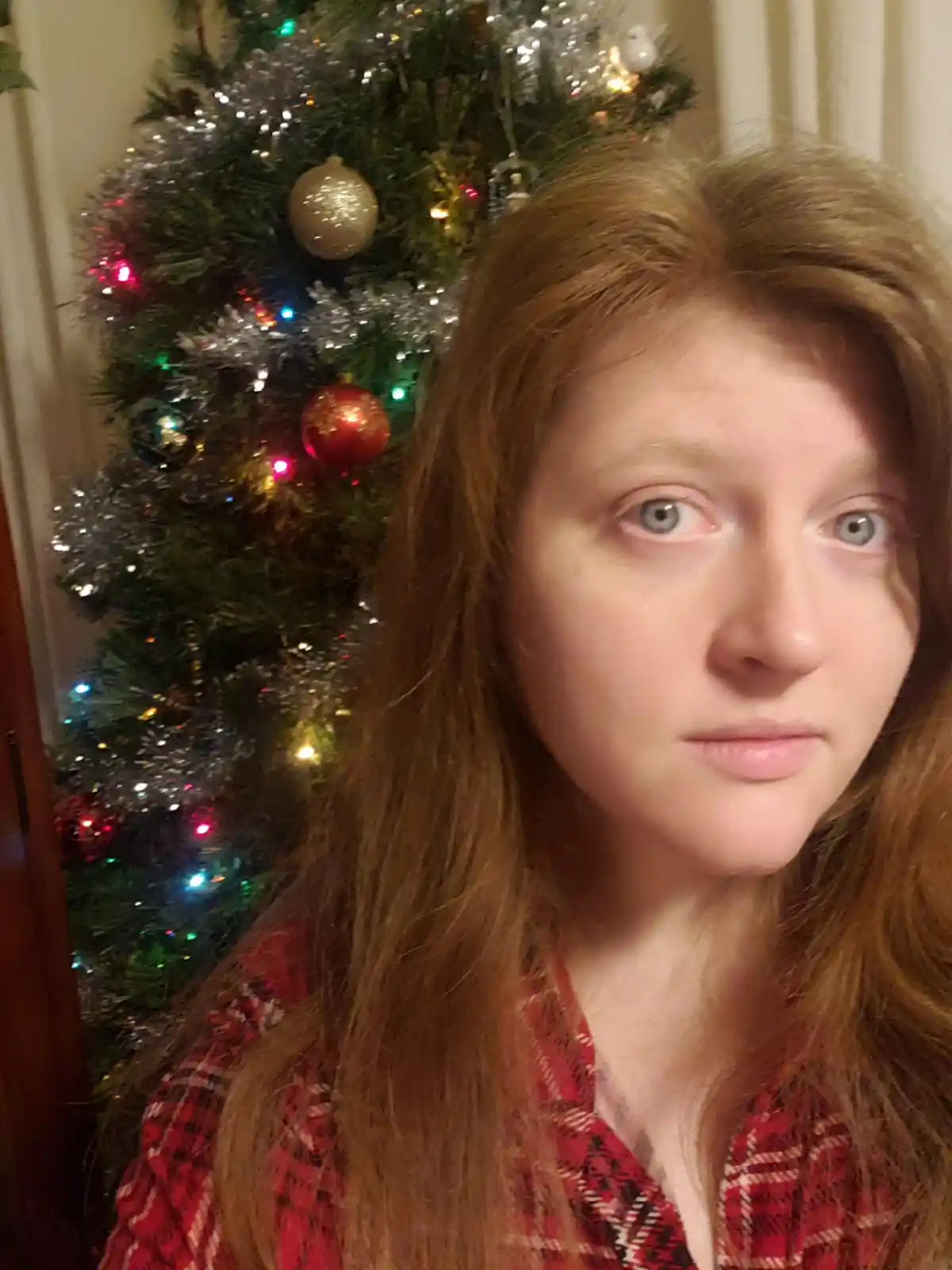 A person with long reddish-brown hair and a red plaid shirt stares blankly at the camera with a neutral, unimpressed…
