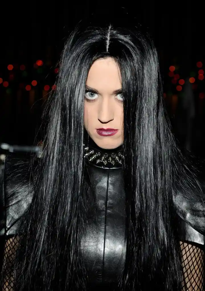 A person with long, straight black hair and pale skin stares intensely at the camera, wearing a black leather outfit with a…