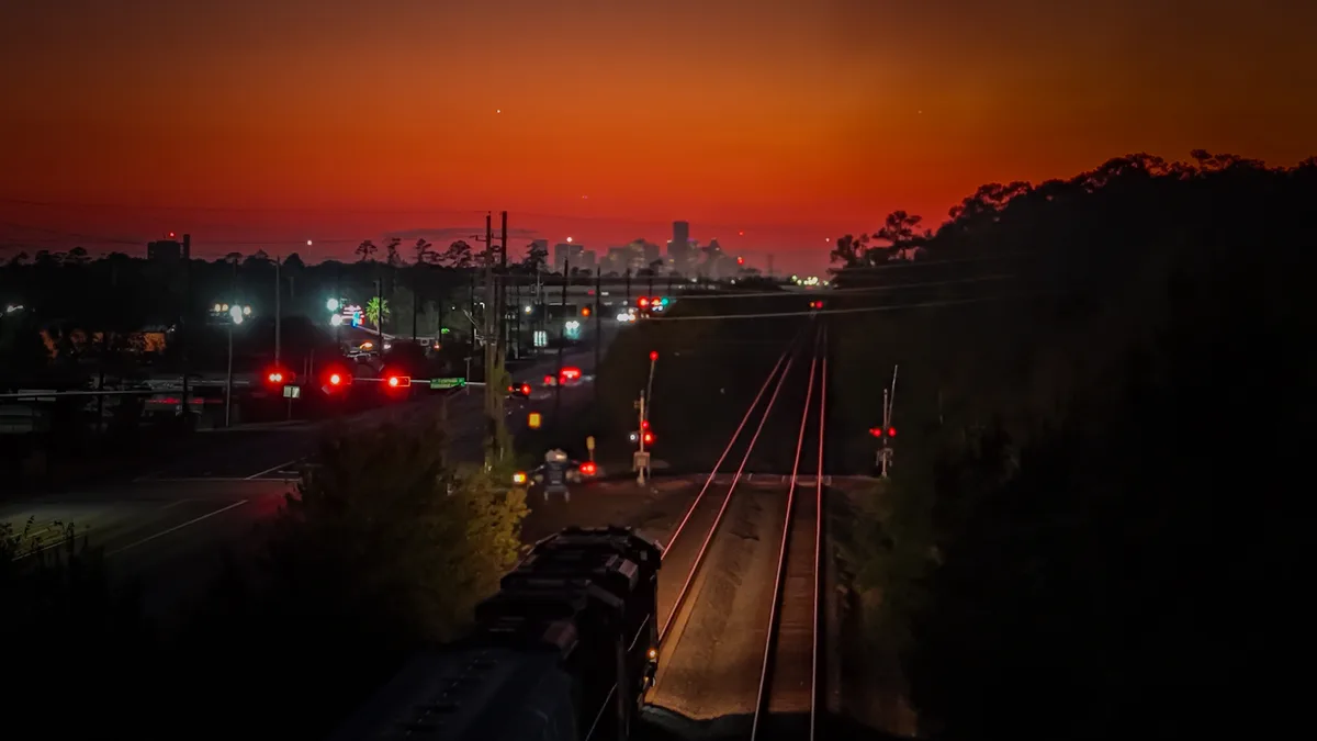 A train is seen from behind, moving along tracks at night under a vivid orange-red sky, with city lights and red traffic…