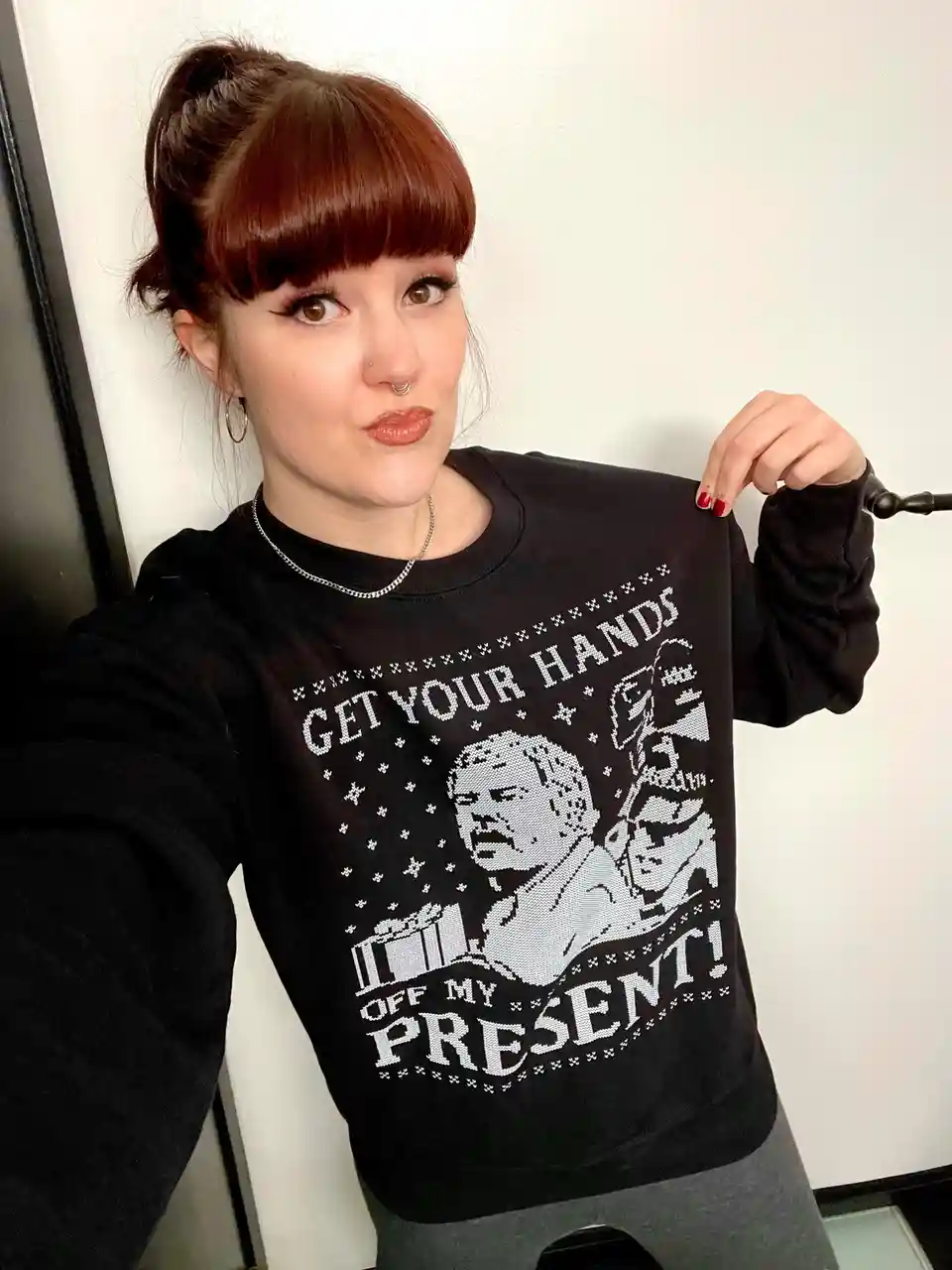 A person with red hair and bangs, wearing a black sweater with a white pixelated graphic, poses while holding up the…