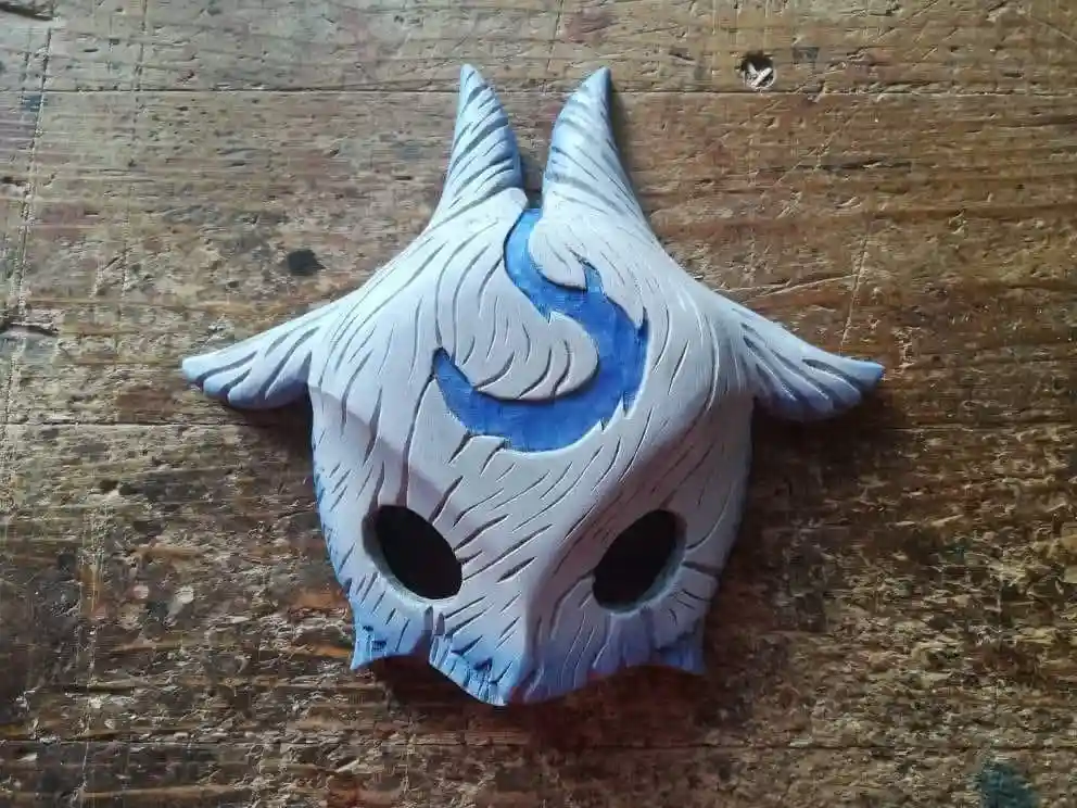 A hand-carved wooden mask with a stylized blue and white design, featuring two pointed horns, wing-like protrusions, and eye…