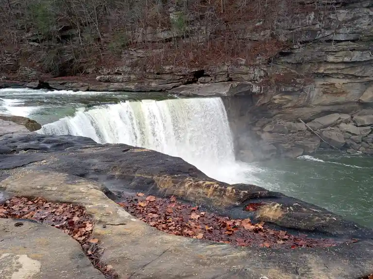 A wide shot of a natural waterfall cascading over dark, wet rocks in a forested area with bare trees and scattered brown…