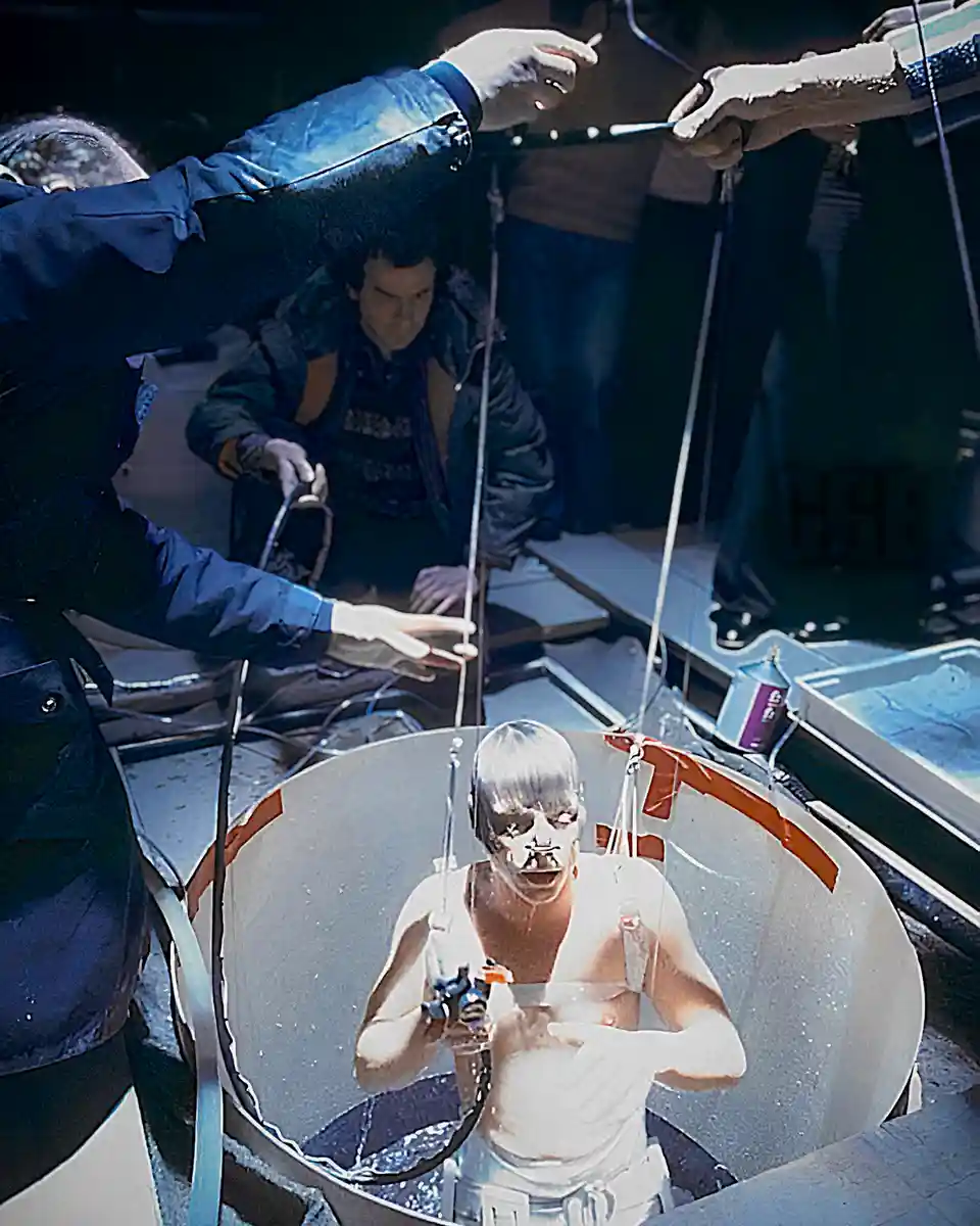 A person with a shaved head and white face paint is suspended in a large, transparent, cone-shaped apparatus, holding a…