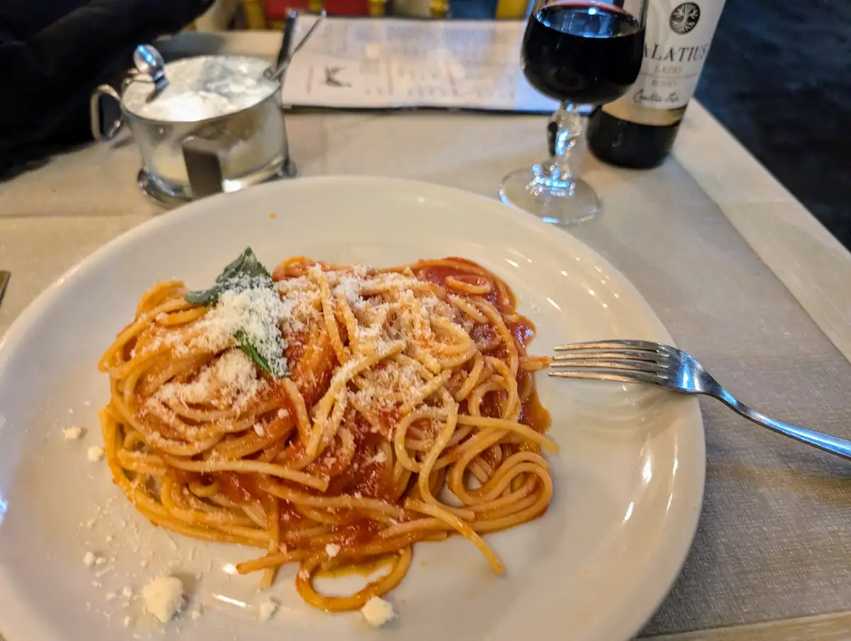 A plate of spaghetti with tomato sauce and grated cheese sits on a restaurant table, with a fork resting beside it. In the…