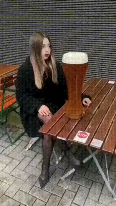 A woman sits at an outdoor wooden table with a giant beer glass. She lifts it to her lips, drinks it all, and then spits out…