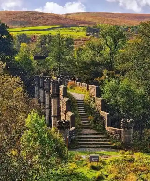 A scenic view of a stone staircase flanked by castle-like stone walls leading up a hillside through green trees, with…