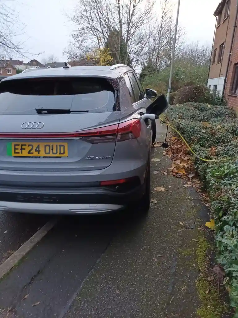 A gray Audi e-tron SUV is parked on a wet residential street, plugged into a charging station with a yellow cable running…