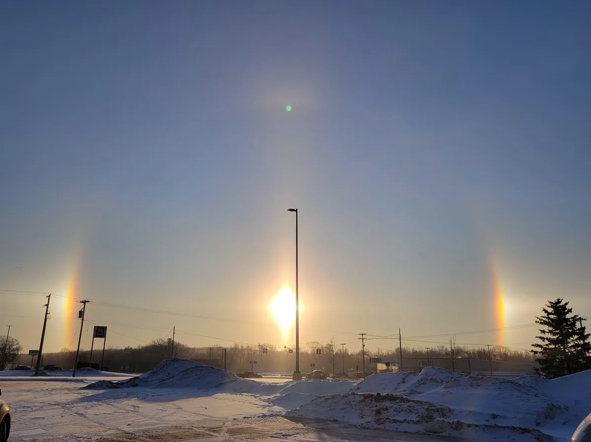 A snowy parking lot at sunrise or sunset with the sun directly behind a tall streetlight, casting a bright glare. Two faint…
