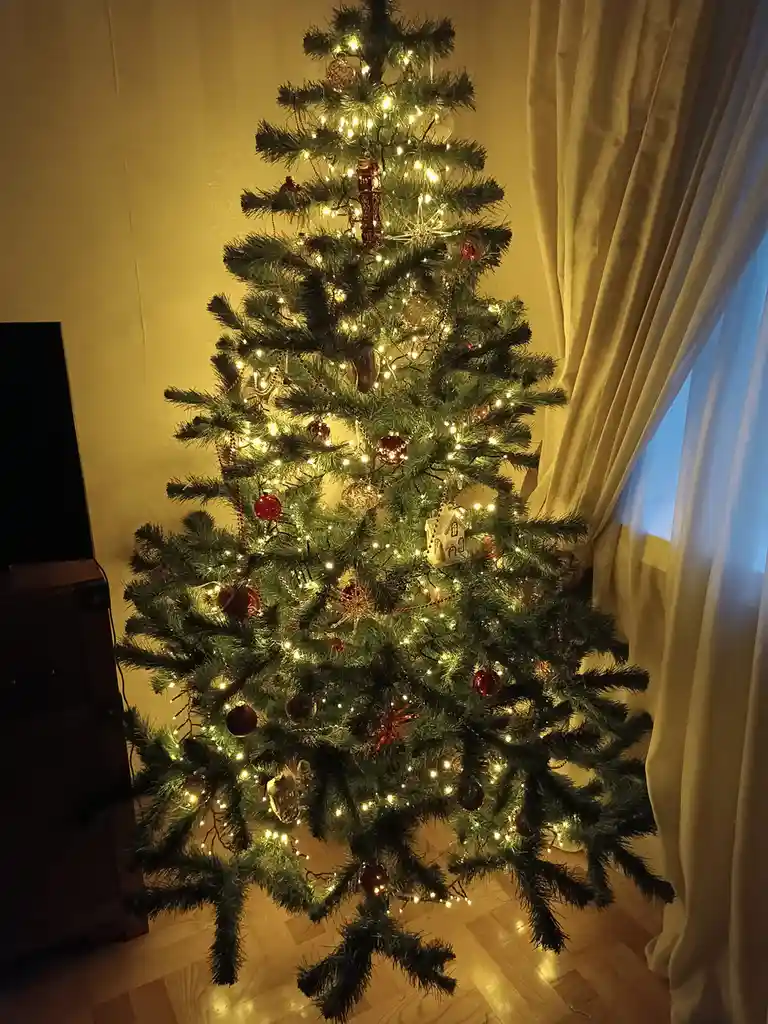 A warmly lit, decorated artificial Christmas tree stands indoors, adorned with string lights, red and gold ornaments, and a…