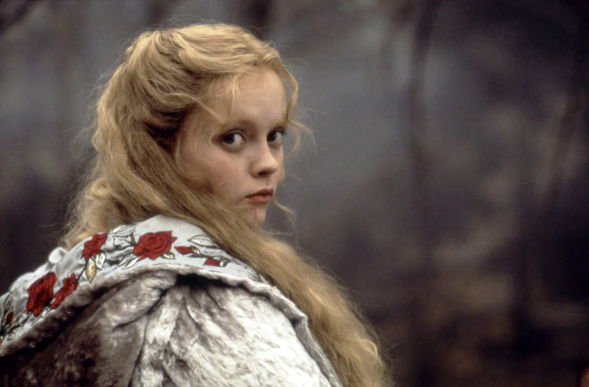 A young girl with long blonde hair, wearing a white coat with red rose embroidery, turns her head to look over her shoulder…