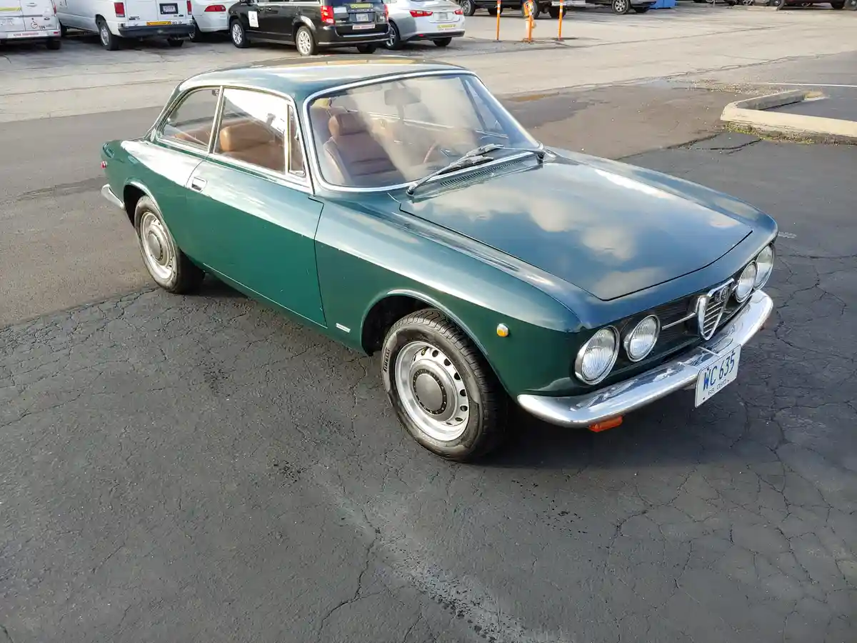 A green vintage Alfa Romeo 2600 coupe is parked in a cracked asphalt lot, its chrome trim and four-headlight grille gleaming…