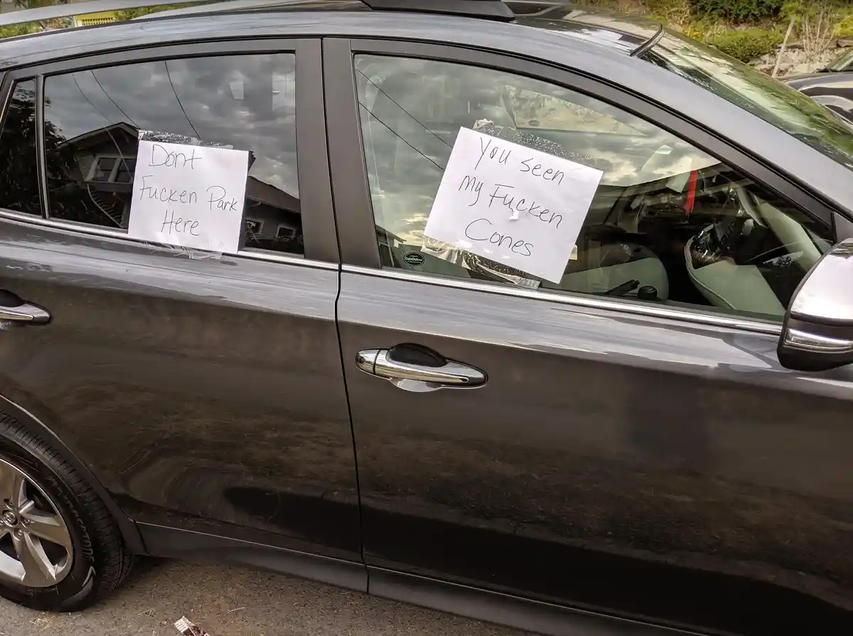 A dark gray car has two handwritten signs taped to its side windows. The left sign reads 'Dont Fucken Park Here' and the…