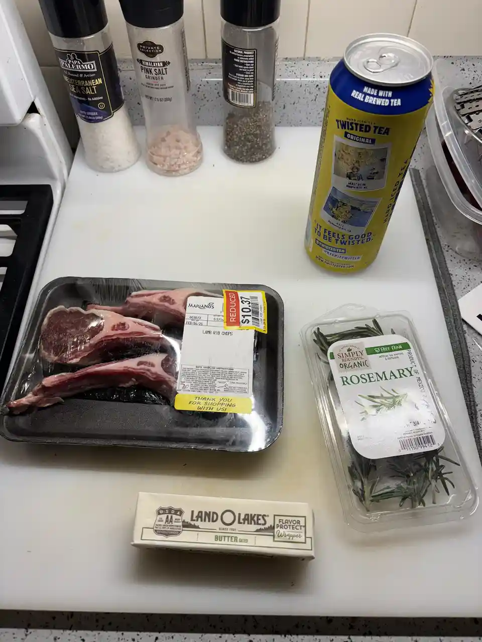A kitchen counter displays ingredients for cooking: lamb rib chops, a package of rosemary, a stick of Land O'Lakes salted…