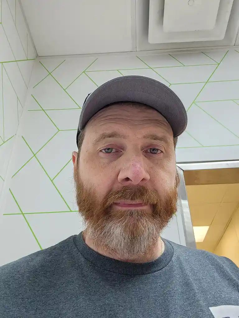 A man with a reddish-brown beard and gray baseball cap stares directly at the camera with a weary expression, standing in…