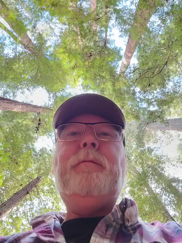 A low-angle selfie shows a man with glasses and a beard, wearing a cap and plaid shirt, looking down at the camera with a…