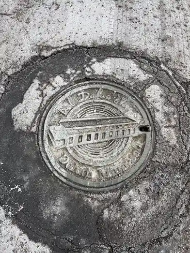 A close-up photo of a weathered, circular metal manhole cover embedded in cracked asphalt. The cover is engraved with 'Bob…