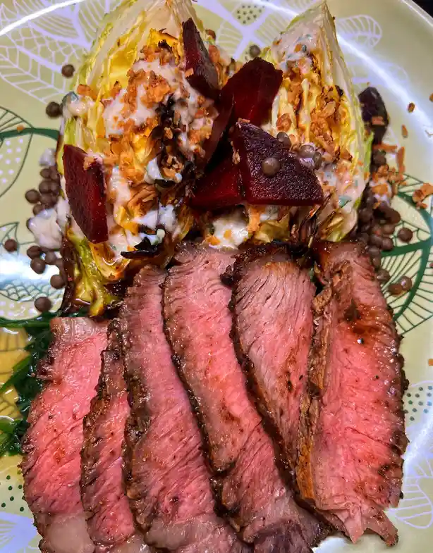 A close-up photo of a plated meal featuring sliced medium-rare beef in the foreground and a wedge salad topped with…