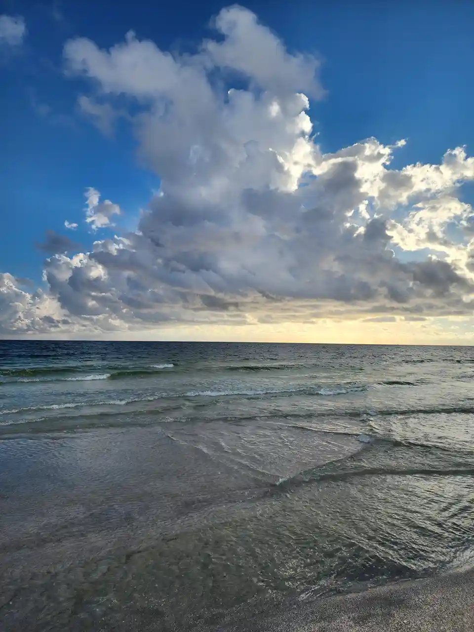 A serene beach scene with gentle waves lapping at the shore under a vast sky filled with dramatic, fluffy clouds illuminated…