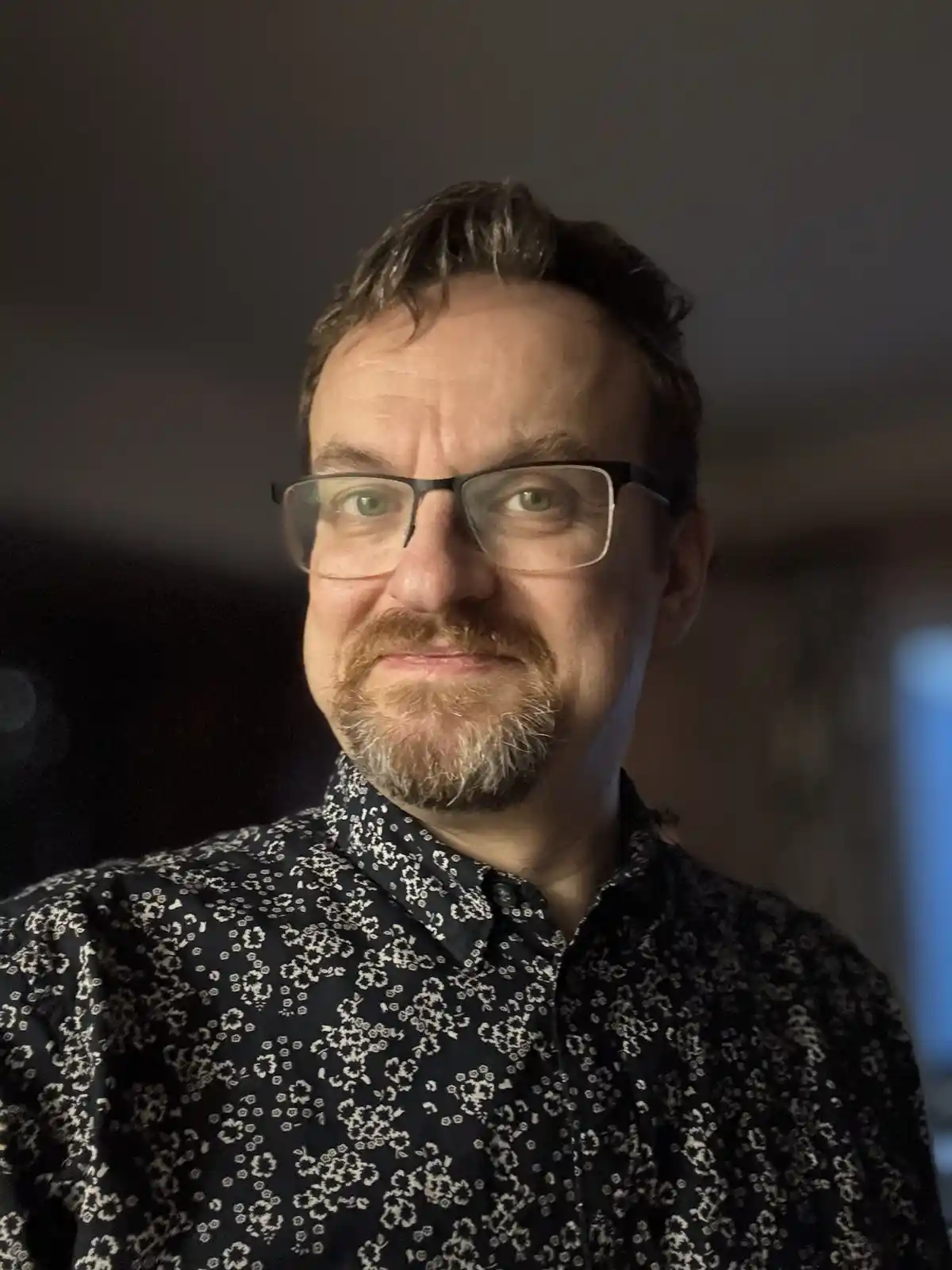 A man with glasses and a goatee, wearing a dark shirt with a white floral pattern, stares directly at the camera with a…