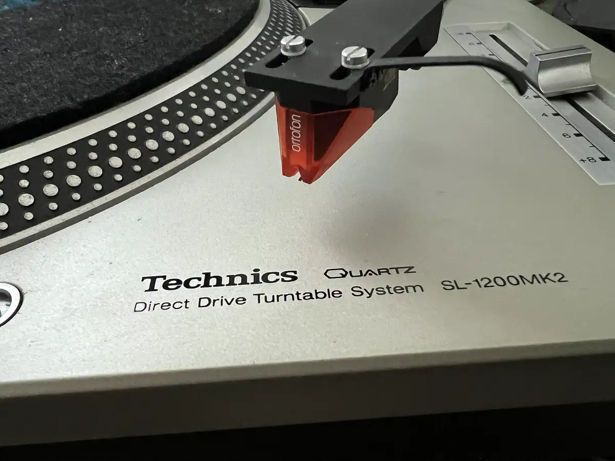 A close-up photo of a Technics SL-1200MK2 turntable, showing its silver body, black platter with white dots, and a red…