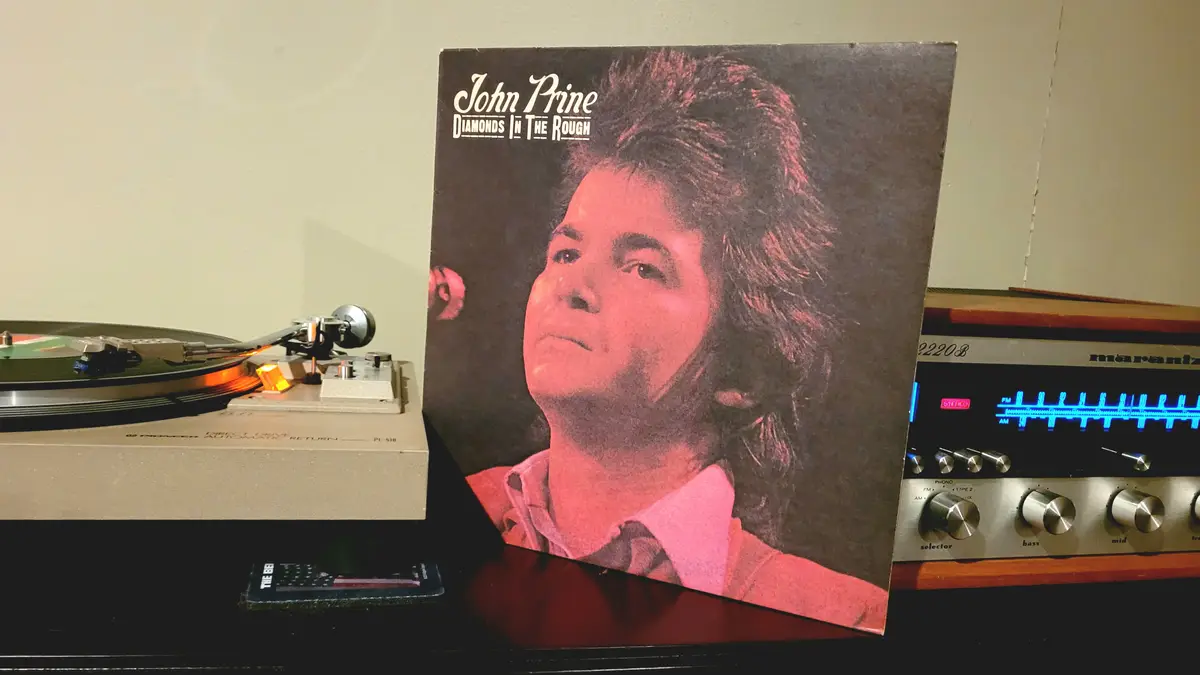 A vintage vinyl record album cover for John Prine's 'Diamonds In The Rough' stands upright on a shelf next to a Pioneer…